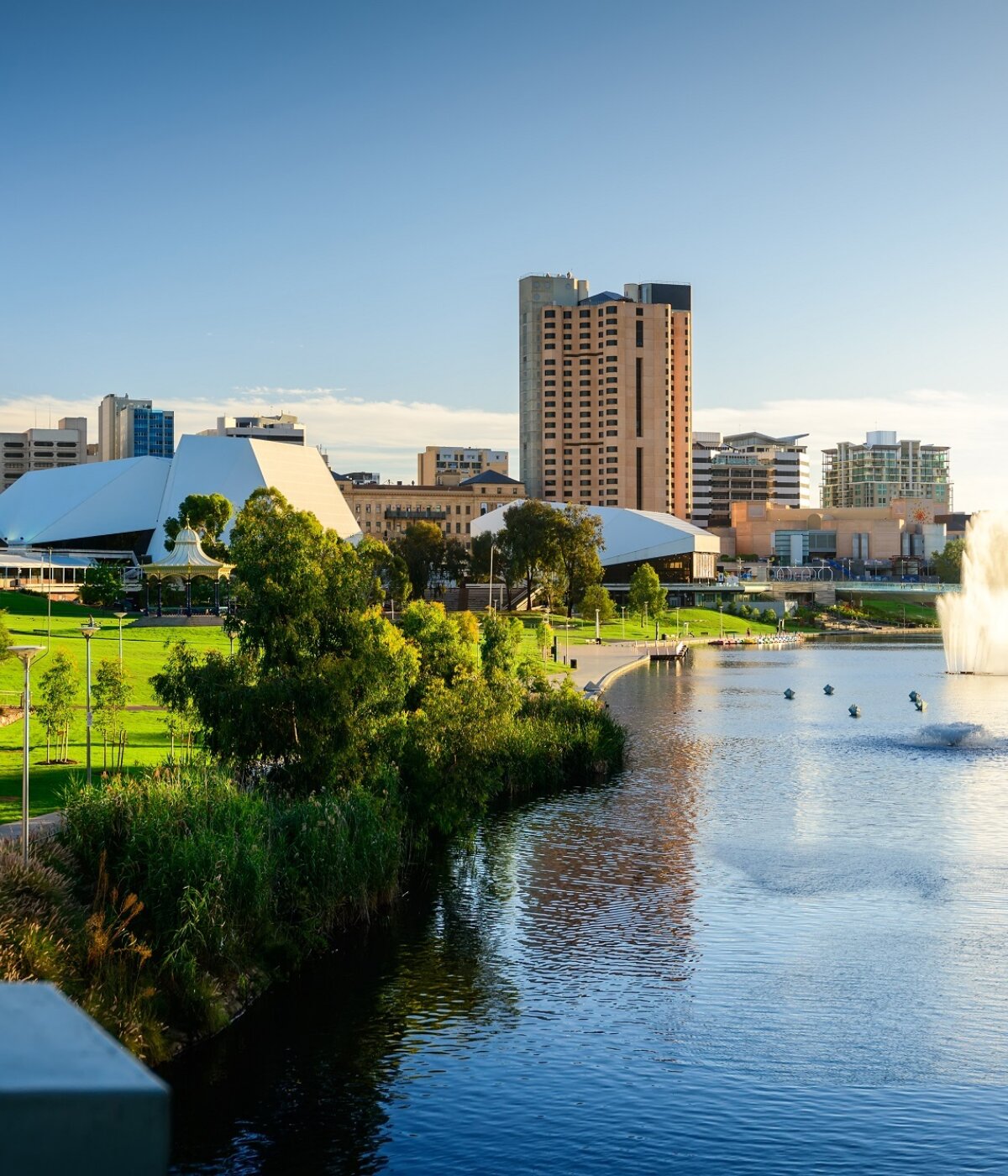 A scenic view of the Adelaide city skyline and riverfront, featuring modern buildings, lush green parks along the riverbank, and a prominent water fountain in the river under a clear blue sky.