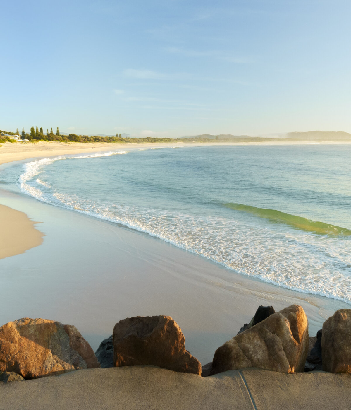 Panoramic view of Blacksmiths Beach with sandy shore, turquoise waves, rocky barrier, trees and clear sky.