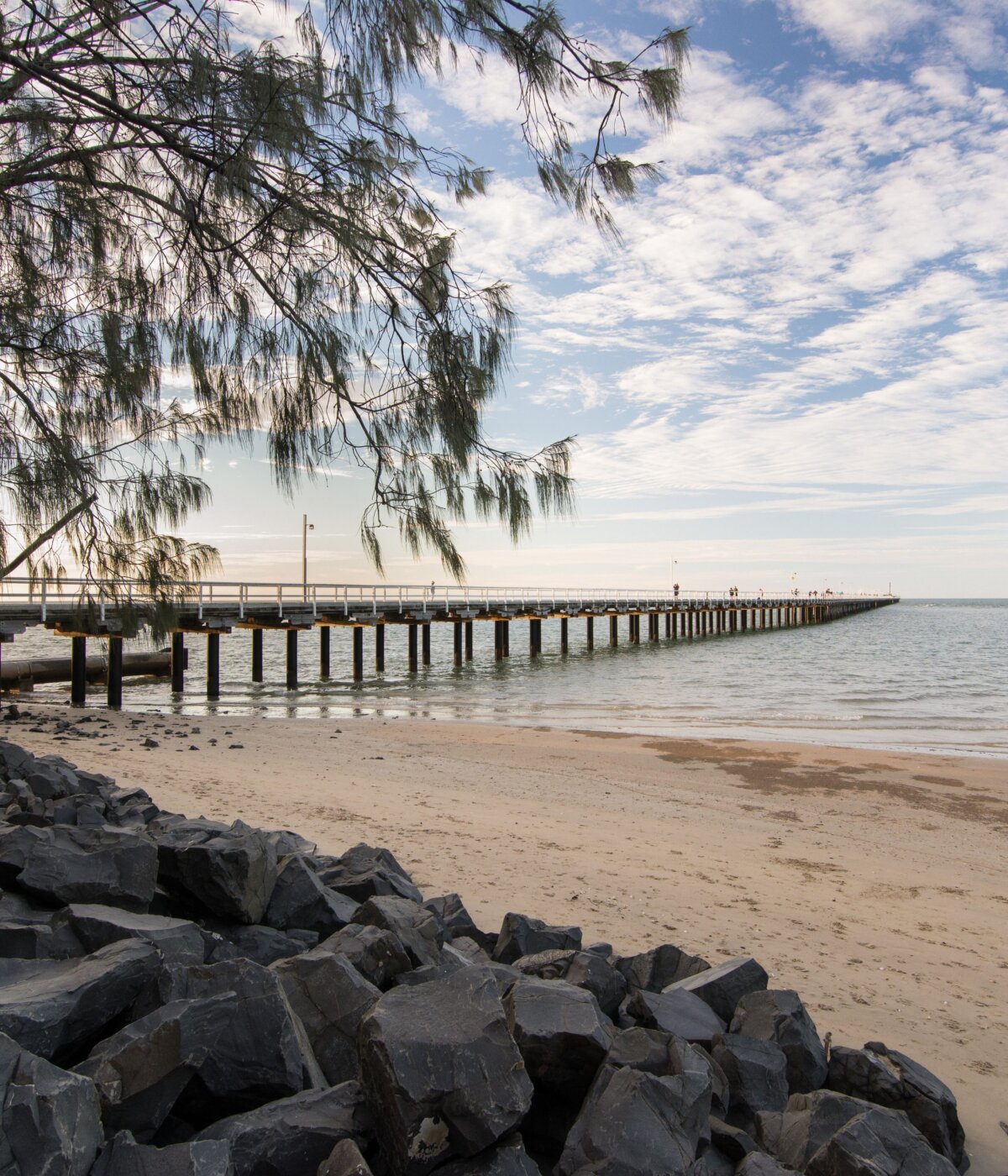 A long pier, identified as Urangan Pier in Hervey Bay, Queensland, Australia, extends into the ocean from a sandy beach with rocks in the foreground. A tree branch with sparse leaves hangs over the scene from the left, and the sky is partly cloudy.