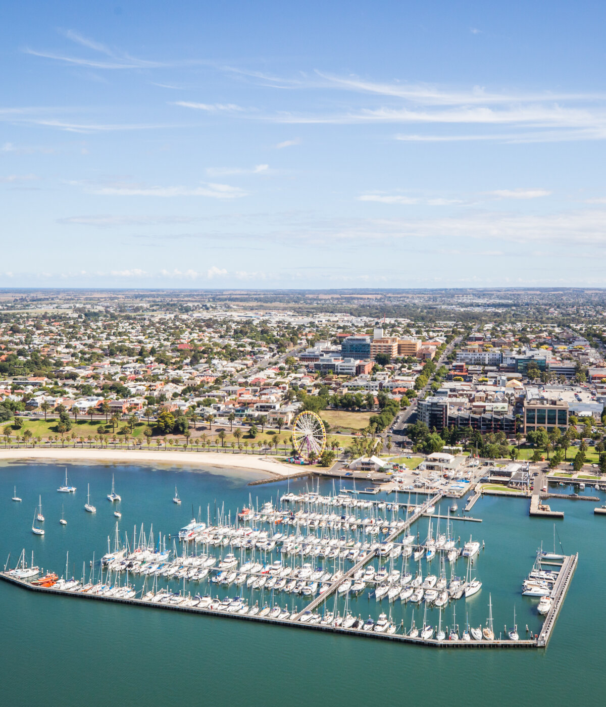 An aerial view of the Geelong waterfront and city, featuring a marina filled with sailboats and yachts, a sandy beach, and urban buildings stretching into the distance under a clear blue sky.