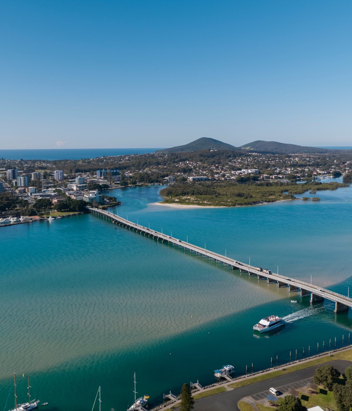 An aerial view of the Forster-Tuncurry Bridge spanning Wallis Lake in Forster, New South Wales, Australia, with boats on the water and a town and hills visible in the background under a clear sky.