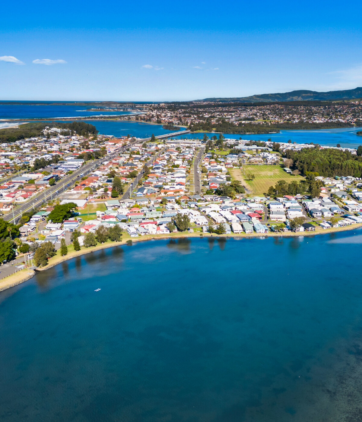 Aerial view of a town situated along the shores of Lake Illawara, also visible is Jettys by the Lake Lifestyle Village, showing residential areas, roads, and a body of water extending towards the horizon.