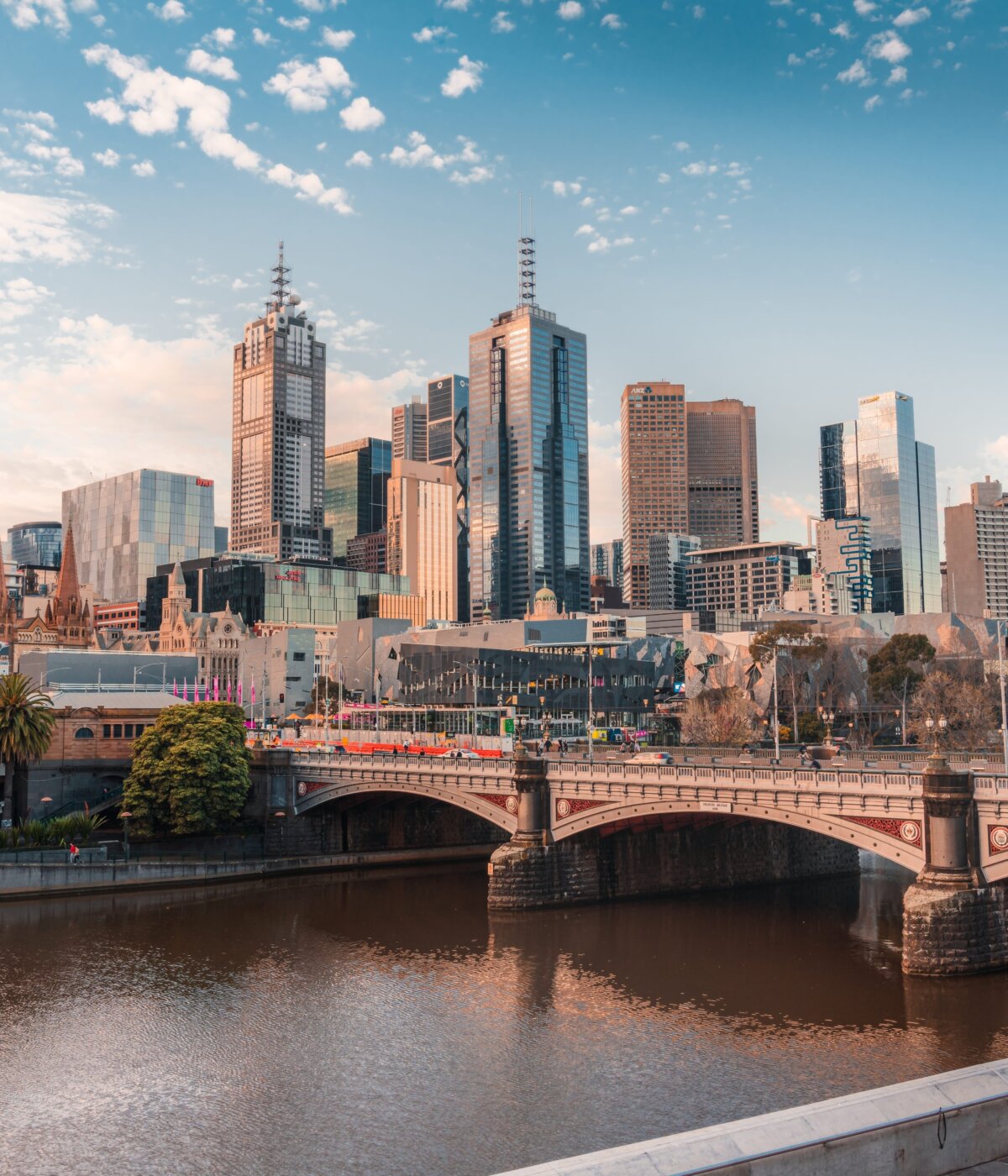 A panoramic view of the Melbourne city skyline at twilight, featuring modern skyscrapers and a historic spire, reflected in the Yarra River with a stone bridge in the foreground, under a partly cloudy sky.