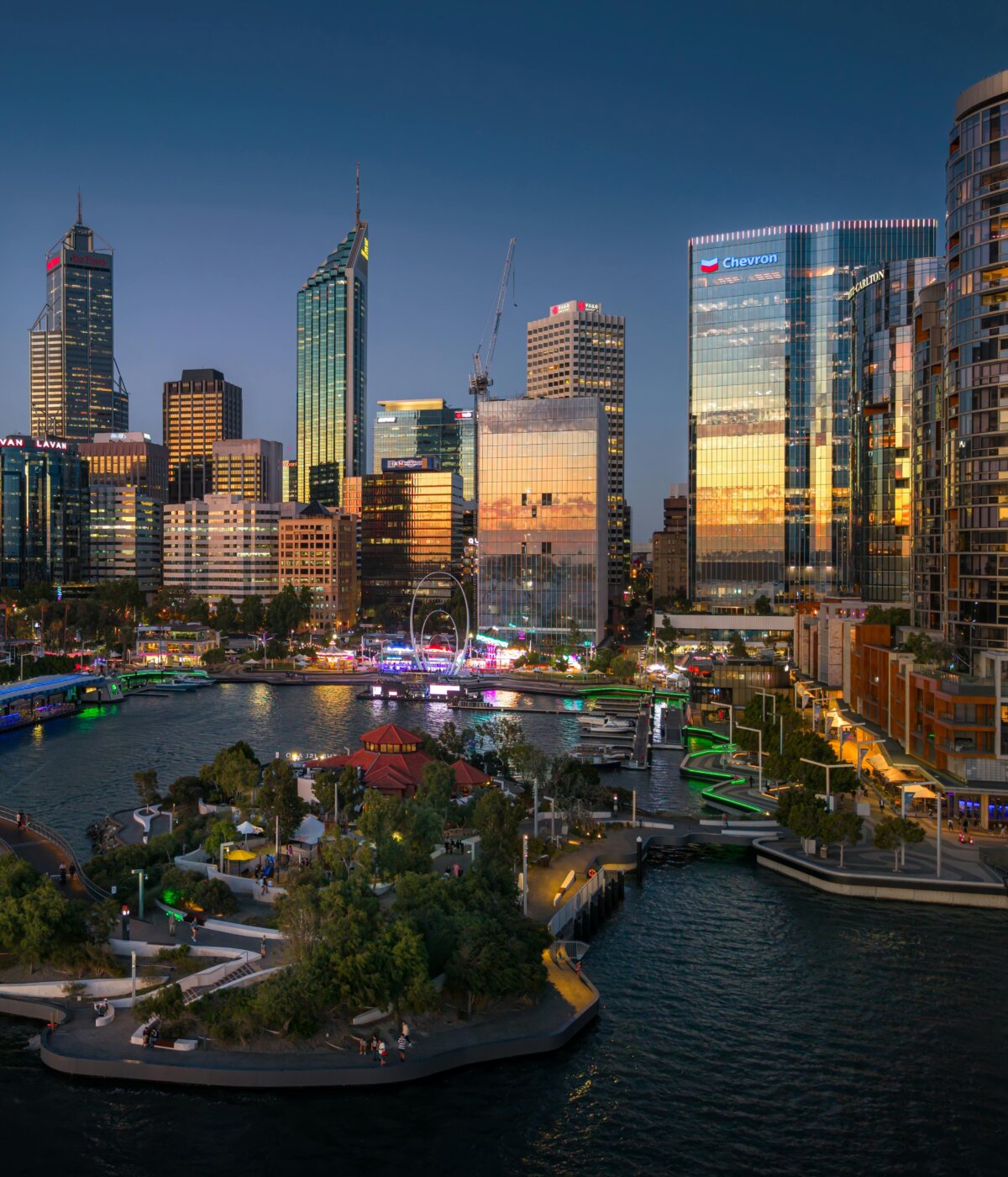 An aerial view of the Perth city skyline at dusk, featuring Elizabeth Quay with its distinctive arched bridge illuminated in green, surrounded by modern skyscrapers and the calm waters reflecting the city lights.