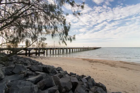 A long pier, identified as Urangan Pier in Hervey Bay, Queensland, Australia, extends into the ocean from a sandy beach with rocks in the foreground. A tree branch with sparse leaves hangs over the scene from the left, and the sky is partly cloudy.