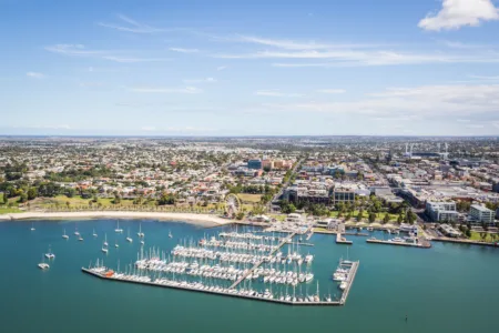 An aerial view of the Geelong waterfront and city, featuring a marina filled with sailboats and yachts, a sandy beach, and urban buildings stretching into the distance under a clear blue sky.