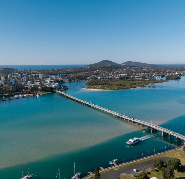 An aerial view of the Forster-Tuncurry Bridge spanning Wallis Lake in Forster, New South Wales, Australia, with boats on the water and a town and hills visible in the background under a clear sky.
