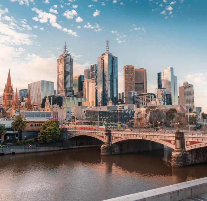 A panoramic view of the Melbourne city skyline at twilight, featuring modern skyscrapers and a historic spire, reflected in the Yarra River with a stone bridge in the foreground, under a partly cloudy sky.