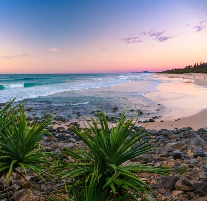 A scenic view of Sunshine Beach in Noosa, Queensland, Australia, at sunset or sunrise, featuring waves crashing on a rocky and sandy shore, with lush coastal vegetation in the foreground and distant hills under a vibrant pink and blue sky.
